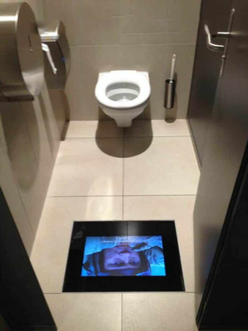 Toilet with a screen on the floor showing a face, in a public restroom.