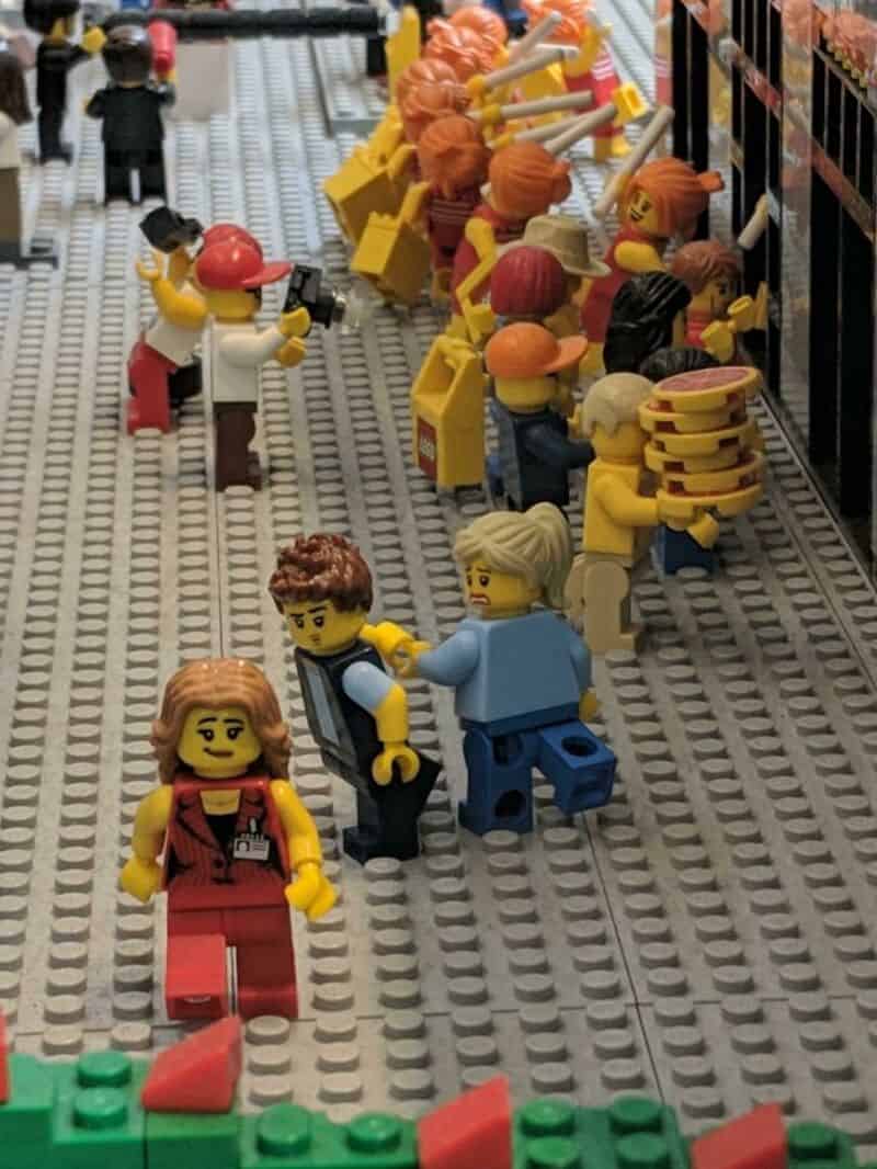 LEGO minifigures shopping in a store aisle with shelves of LEGO products.