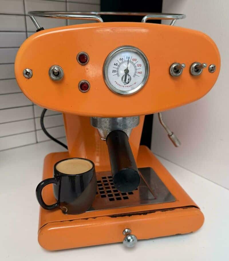 Orange vintage espresso machine with a black mug in front, gauge and controls on top.