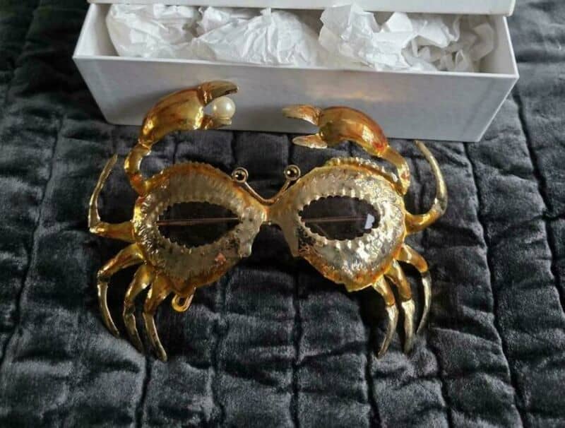 Gold crab-shaped brooch with pearl eyes on black fabric, in a white box.