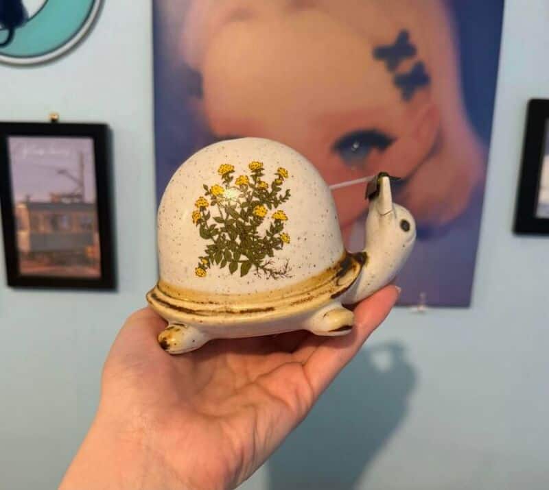 Hand holding a ceramic snail-shaped teapot with yellow flowers design, art gallery background.