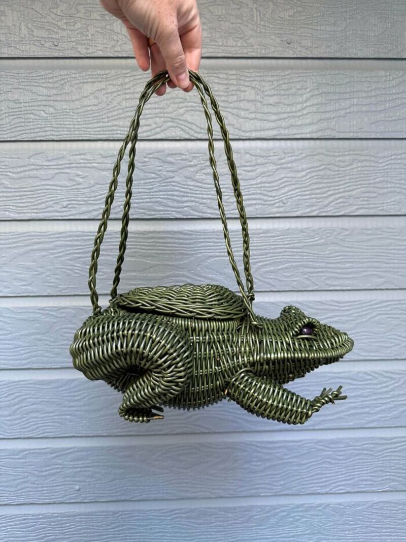 Hand holding a frog-shaped woven bag with a handle, against a gray wooden wall.