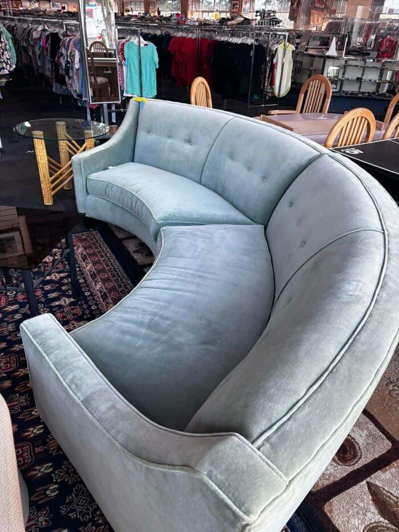 Light blue velvet curved sofa in thrift store with chairs and clothing racks in background.