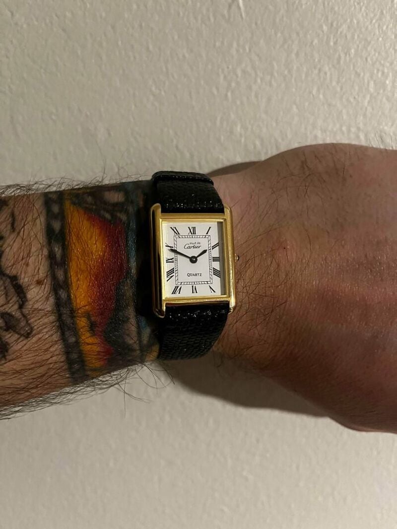 Wristwatch on a person's wrist with a tattoo, against a plain wall.