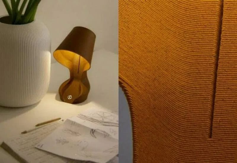 Desk with sketches, a white vase, and a brown lamp, next to a textured orange wall.