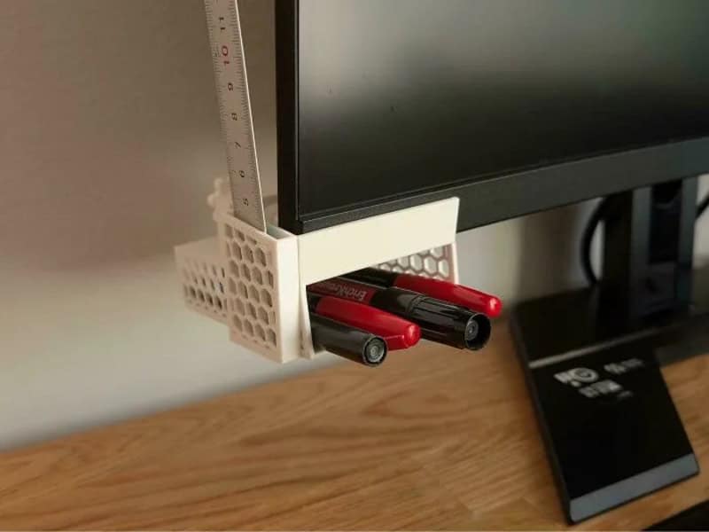 3D-printed monitor stand with pens and ruler attached, on a wooden desk.