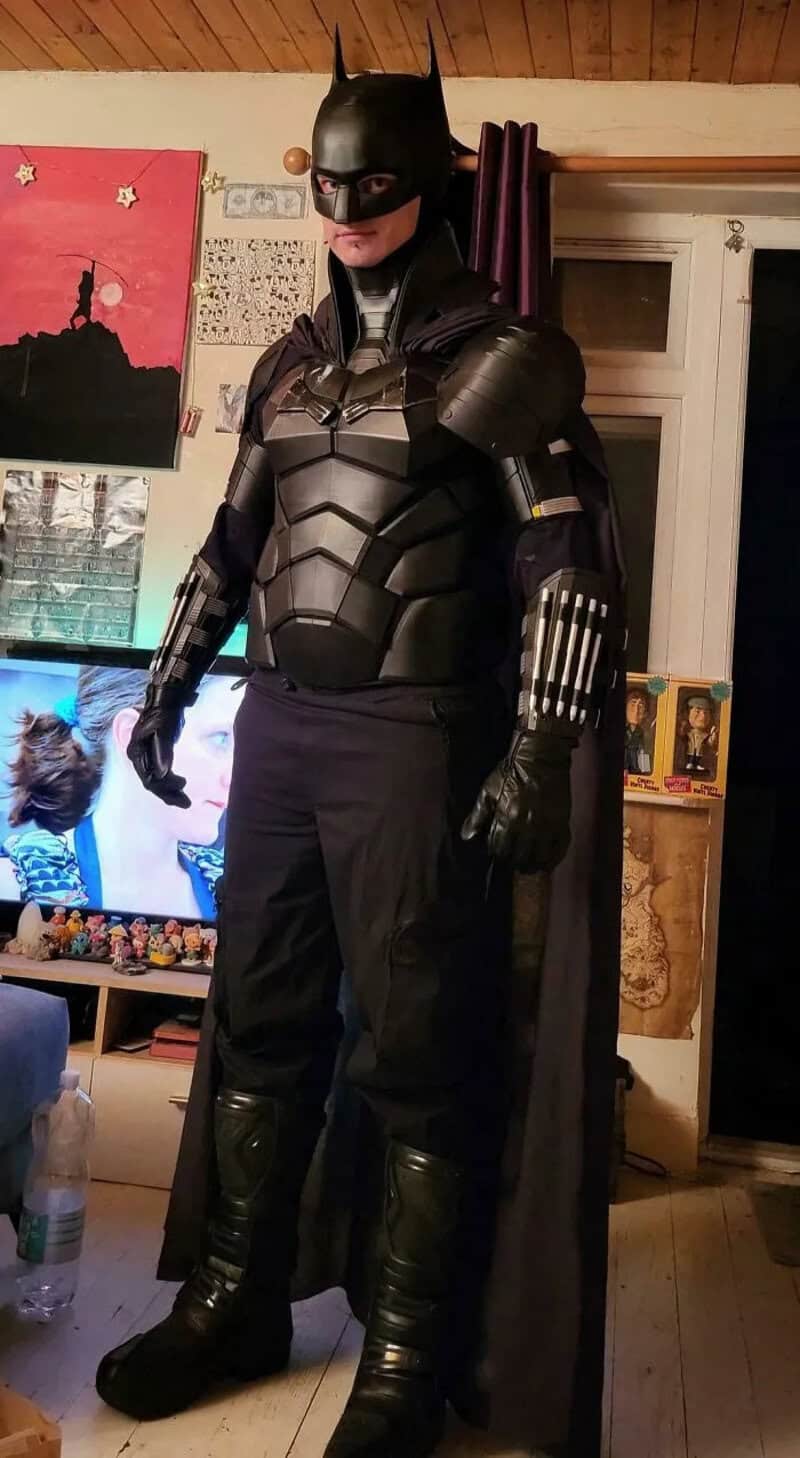 Person dressed as Batman in a costume with a cape, standing indoors.