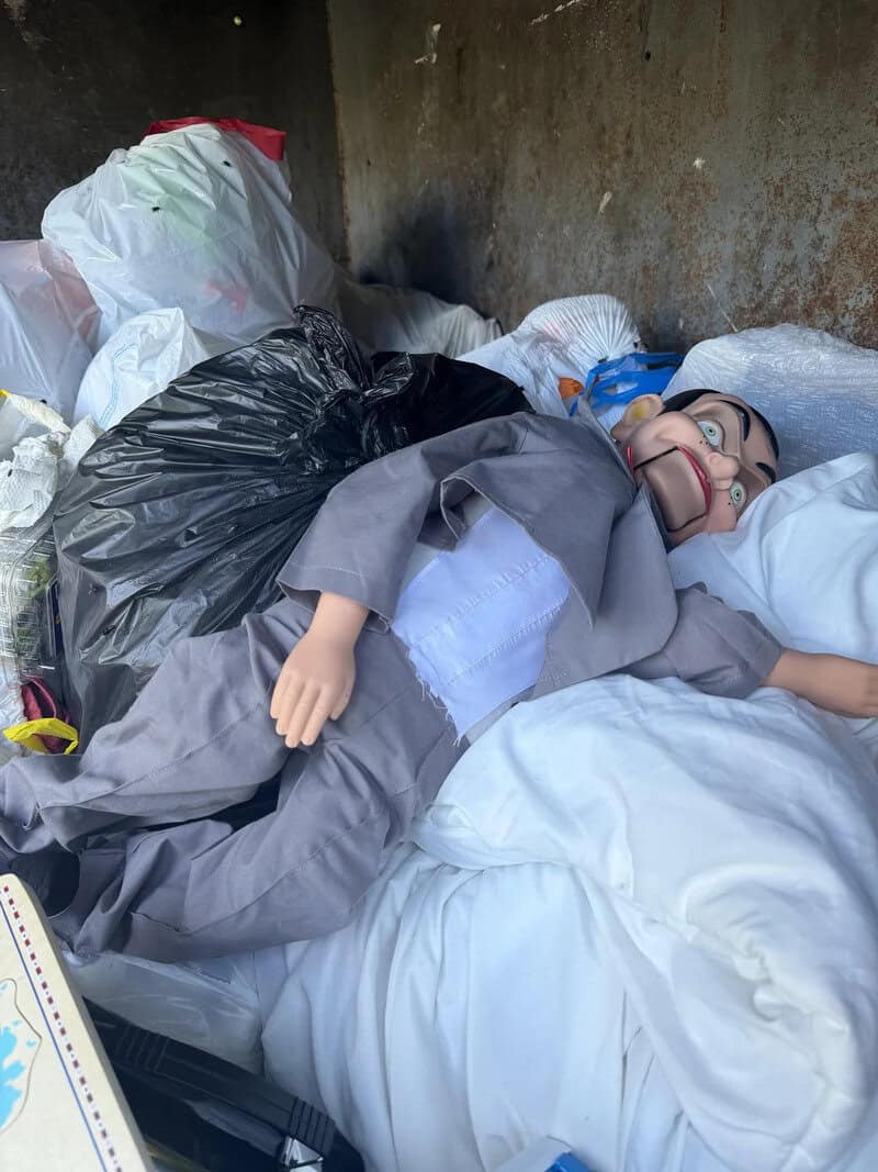 A doll lying on a bed among trash bags and bedding in a dirty room.