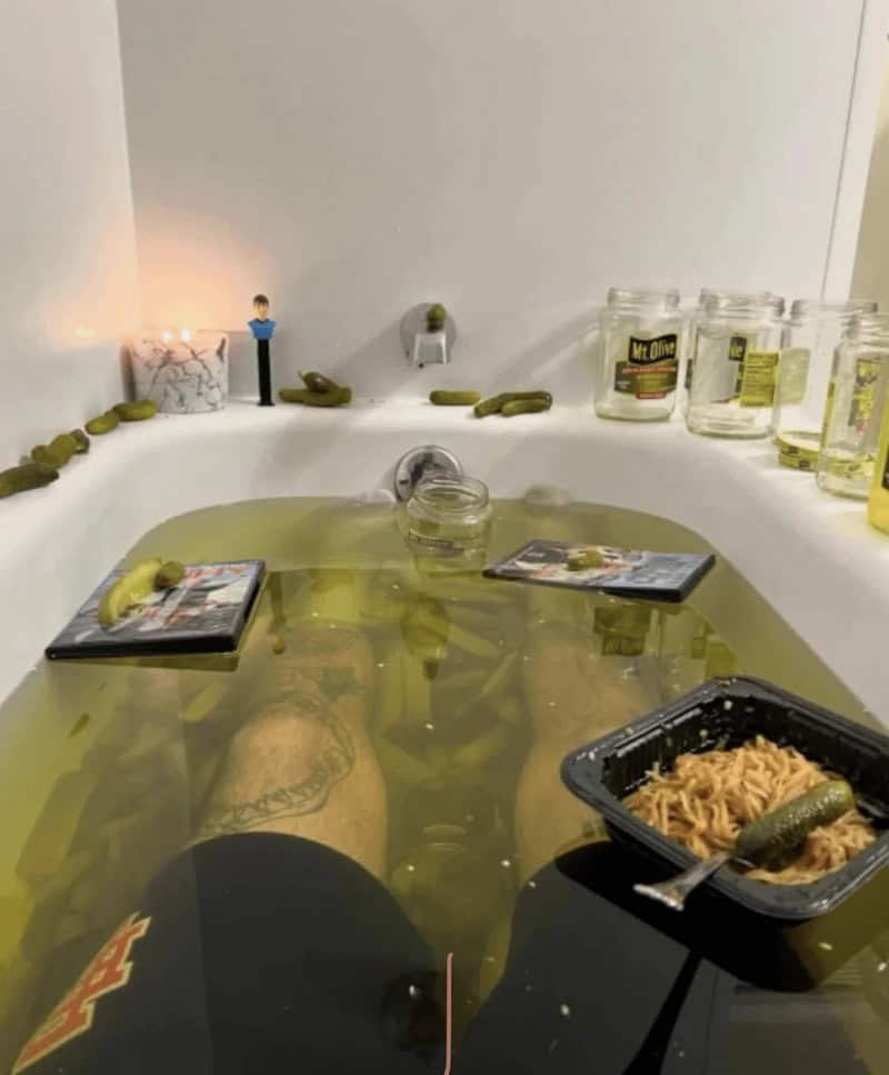 Person soaking in a bathtub filled with yellowish water, surrounded by various items.