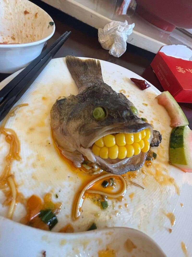 Fish head with corn in mouth on a plate, surrounded by food remnants and chopsticks.