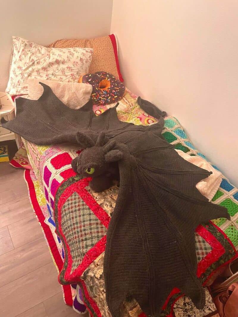 Black dragon plush toy with green eyes and wings on a bed with colorful crochet blankets.