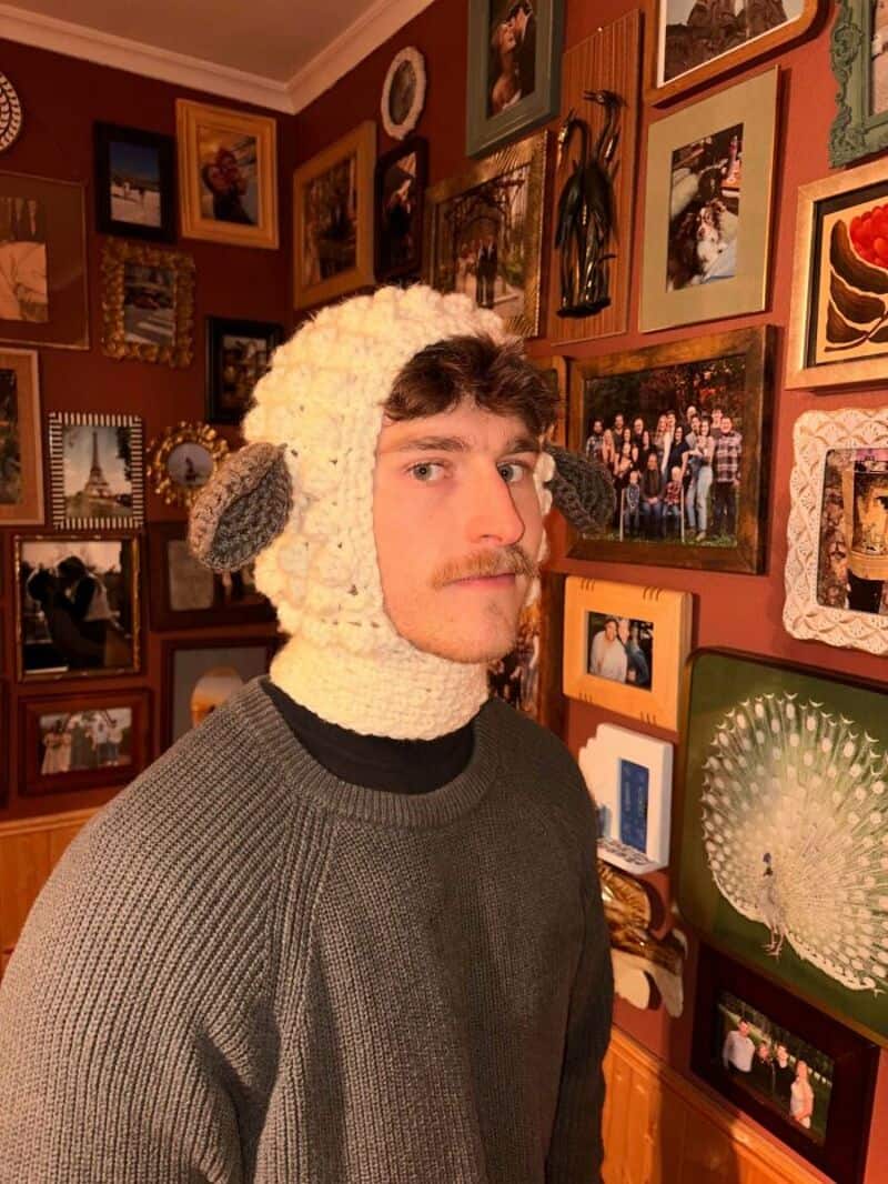 Man wearing a crochet sheep hat in a room with framed photos and art.