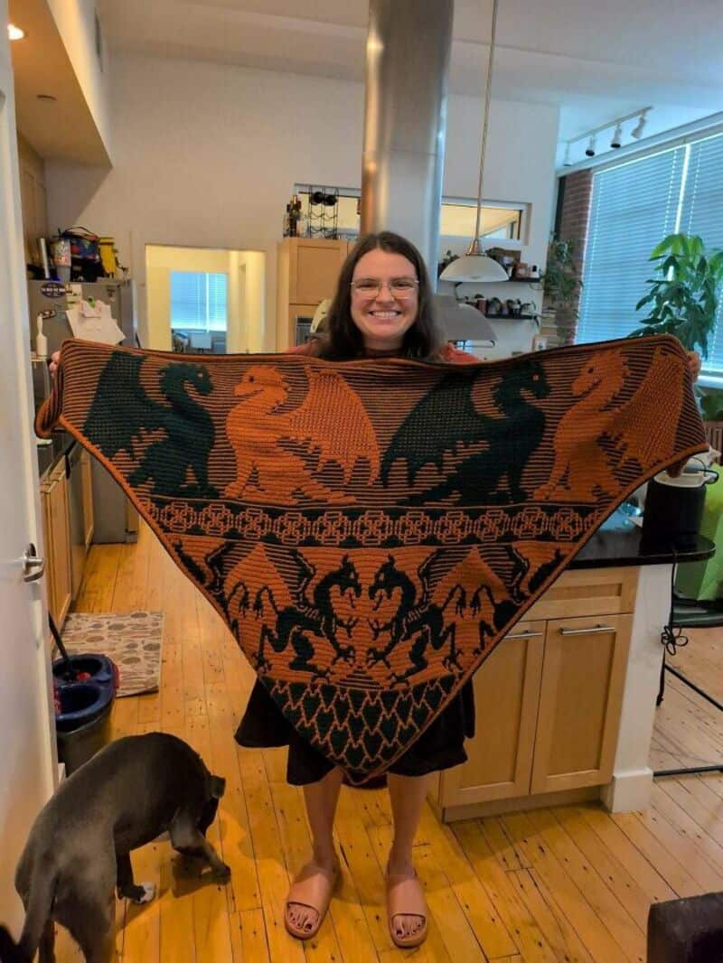 Woman holding a large, colorful crochet blanket with dragon designs in a kitchen.