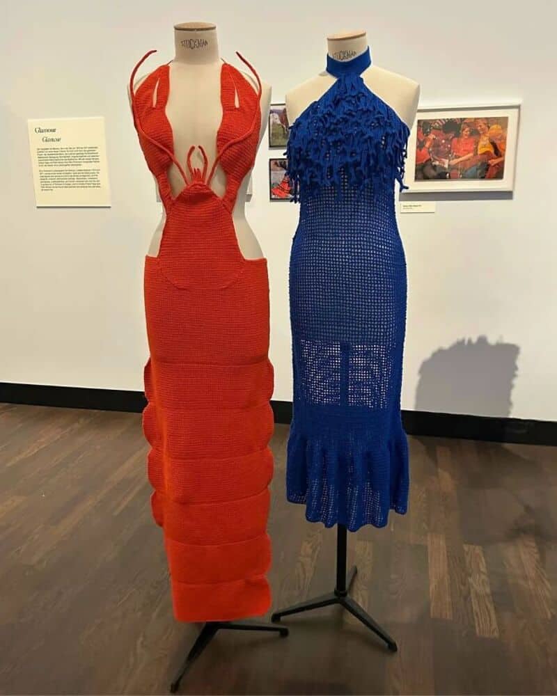 Two mannequins display red and blue crochet dresses in an art gallery.
