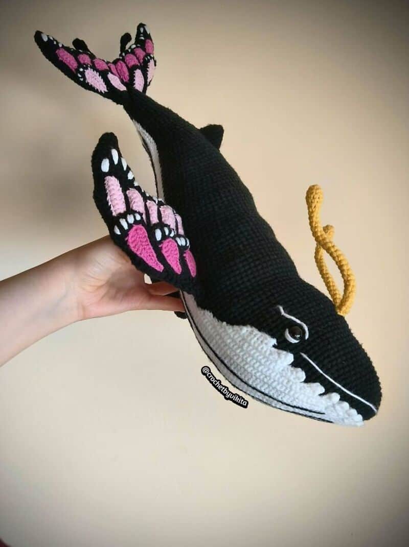 Crochet whale with pink and black butterfly wings, held in hand.
