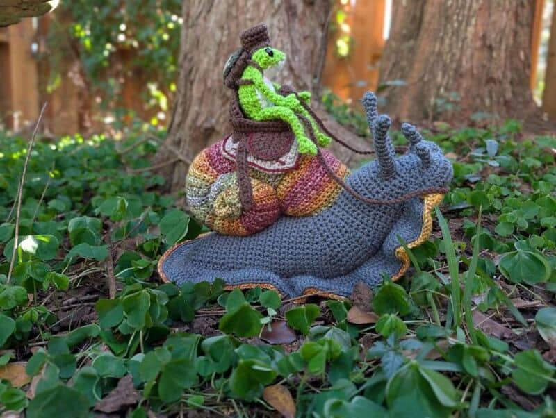 Crochet snail with a green frog riding on its back in a garden.