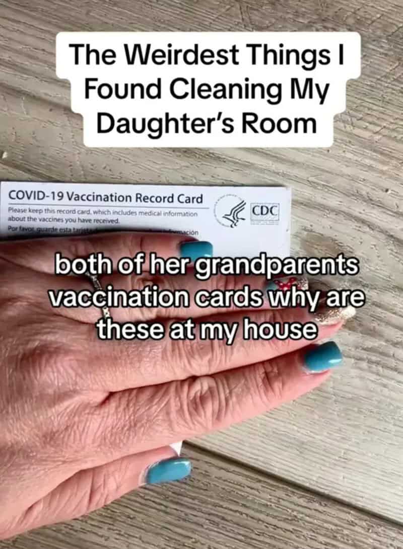The image shows a hand with blue nail polish holding two vaccination cards on a wooden surface.