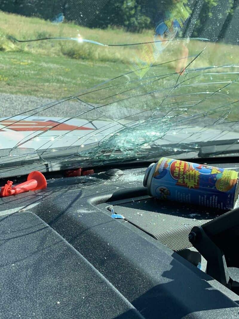 Shattered windshield with a can and toy on dashboard, outside trees visible.