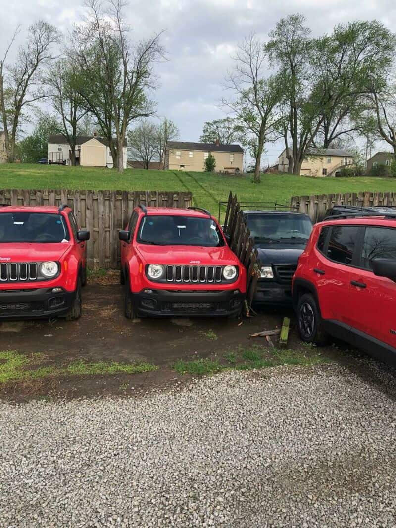 Three red cars parked in a lot with a broken fence and a black car between two of them.
