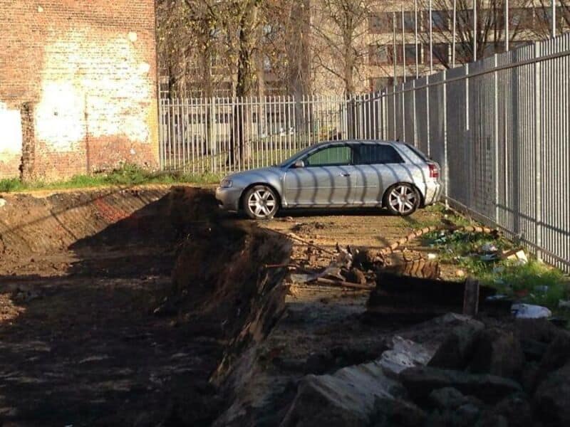 Silver car parked on a dirt patch near a fence, with a damaged ground surface.