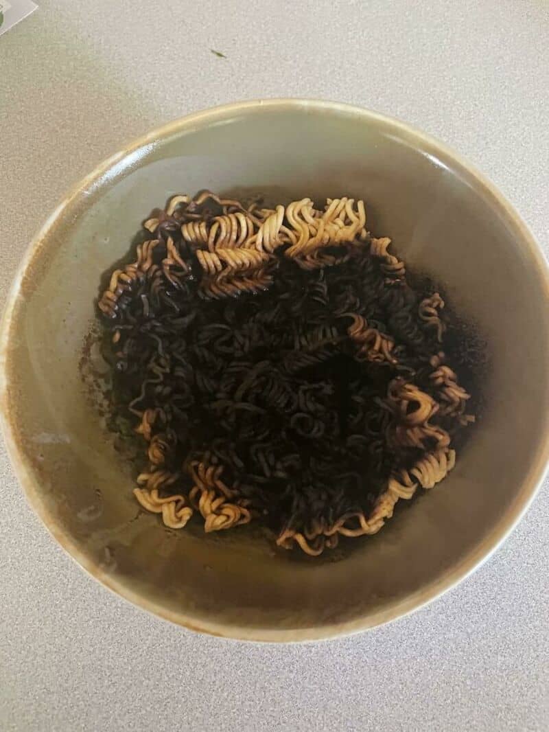 Bowl of cooked instant noodles with dark soy sauce, on a white surface.