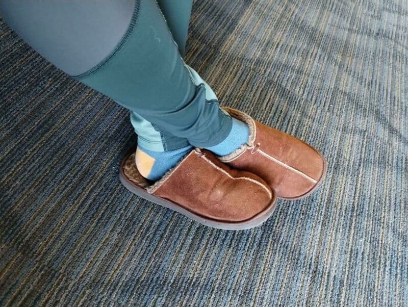 Person wearing brown slippers and blue jeans standing on a carpeted floor.