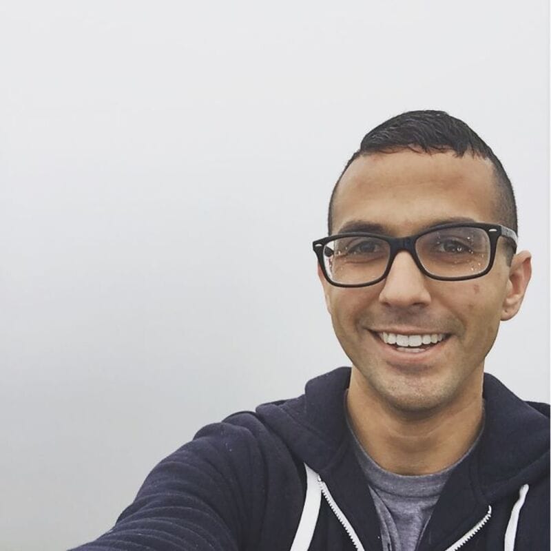 Smiling man with glasses taking a selfie against a plain white background.