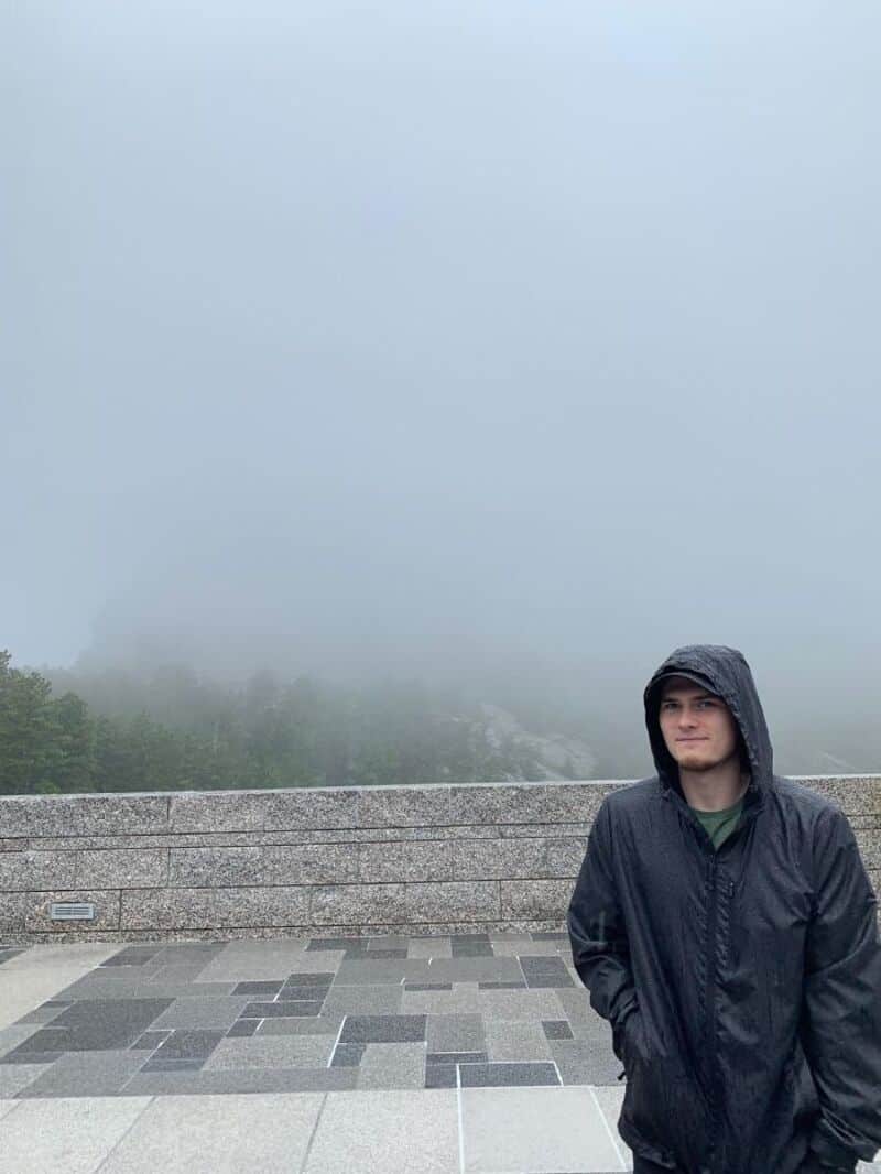 Young man in black raincoat on foggy day at scenic overlook.