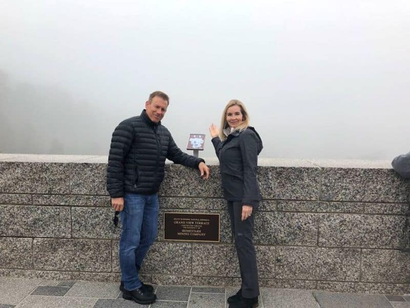 Two people stand by a foggy view with a plaque on a stone wall.