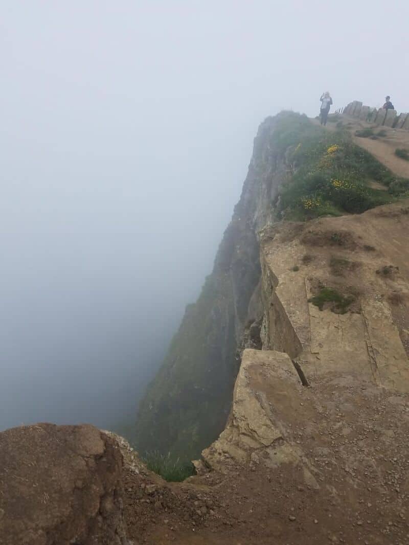 Cliff edge with fog and two hikers standing at the top, overlooking the drop.