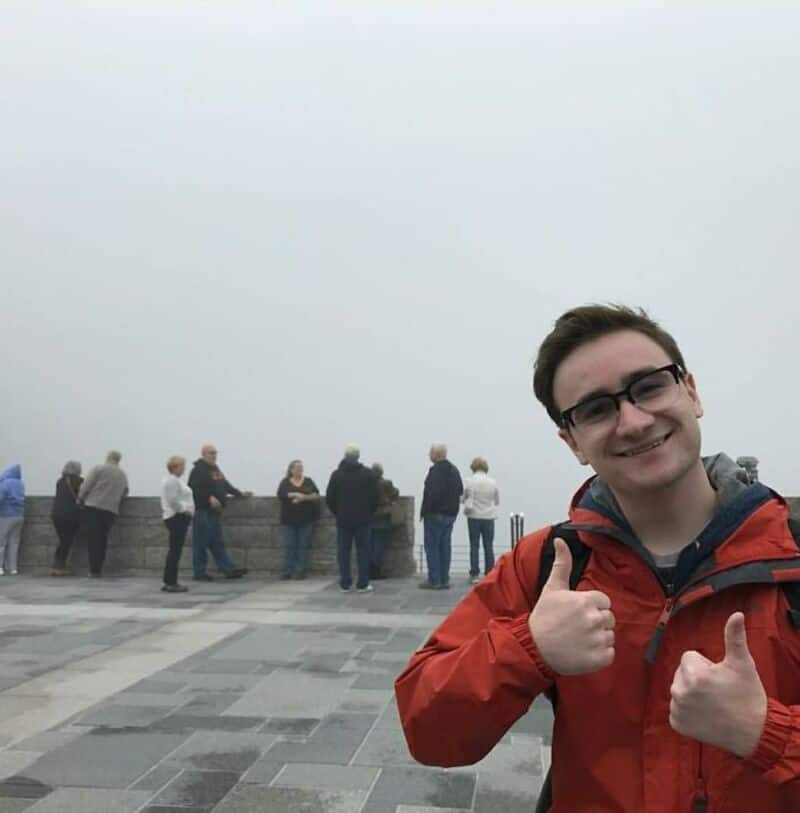 Young man in red jacket giving thumbs up at a foggy viewpoint with people in background.