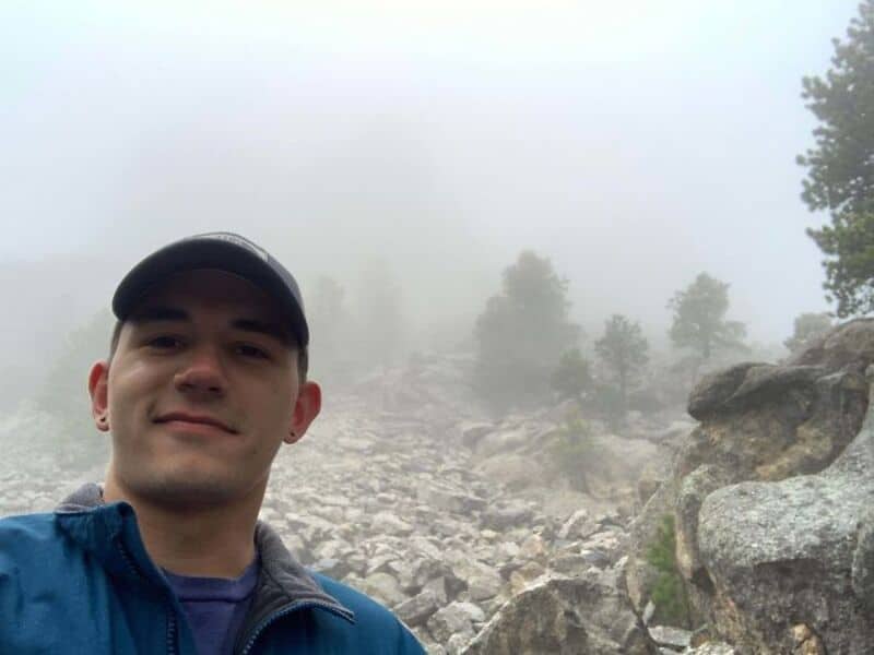 Young man in a blue jacket and black cap taking a selfie in a foggy rocky landscape.