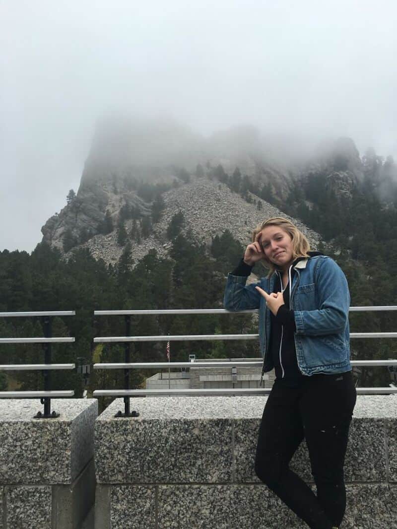 Young woman in denim jacket making a peace sign in front of misty mountains.