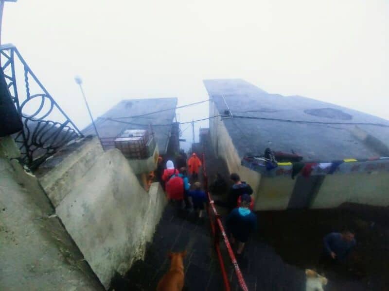 People walking on a narrow path between two large concrete structures in foggy weather.