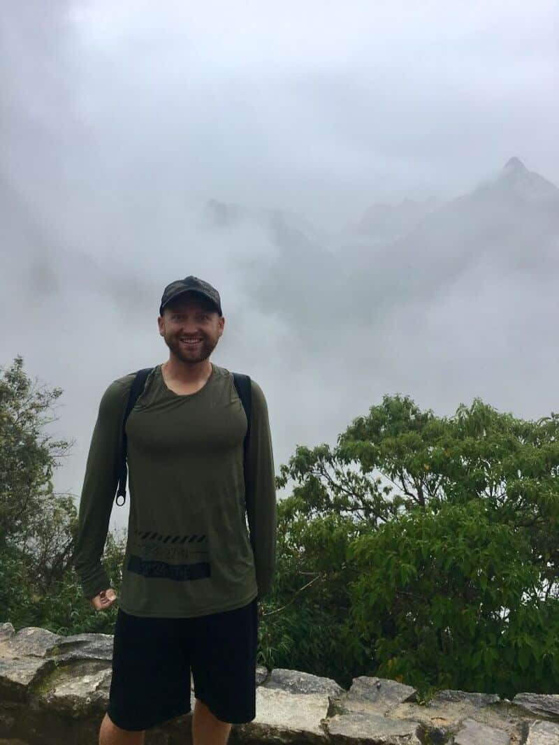 Man in green shirt and black shorts on a foggy mountain trail with lush greenery.