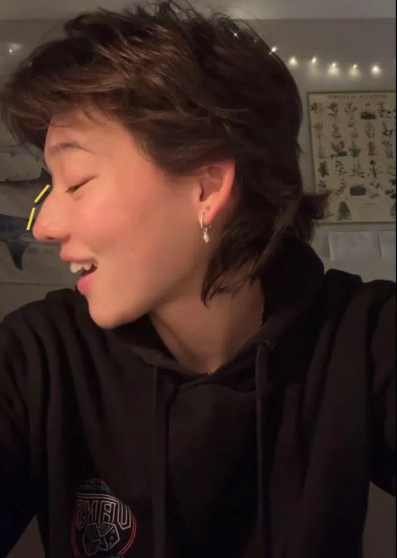 Young person with short brown hair smiling, wearing a black hoodie and hoop earrings.