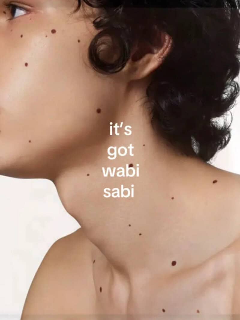 It's got wabi sabi text on a person's neck with multiple moles and curly hair.