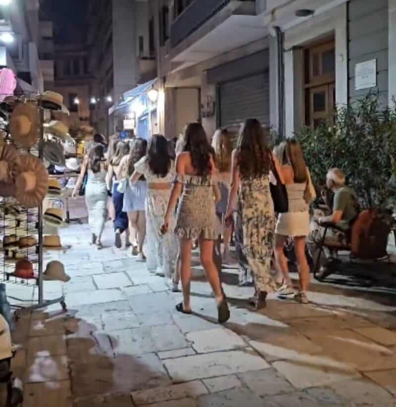 Group of women walking on a city street at night, some wearing dresses, hats on display on the left.