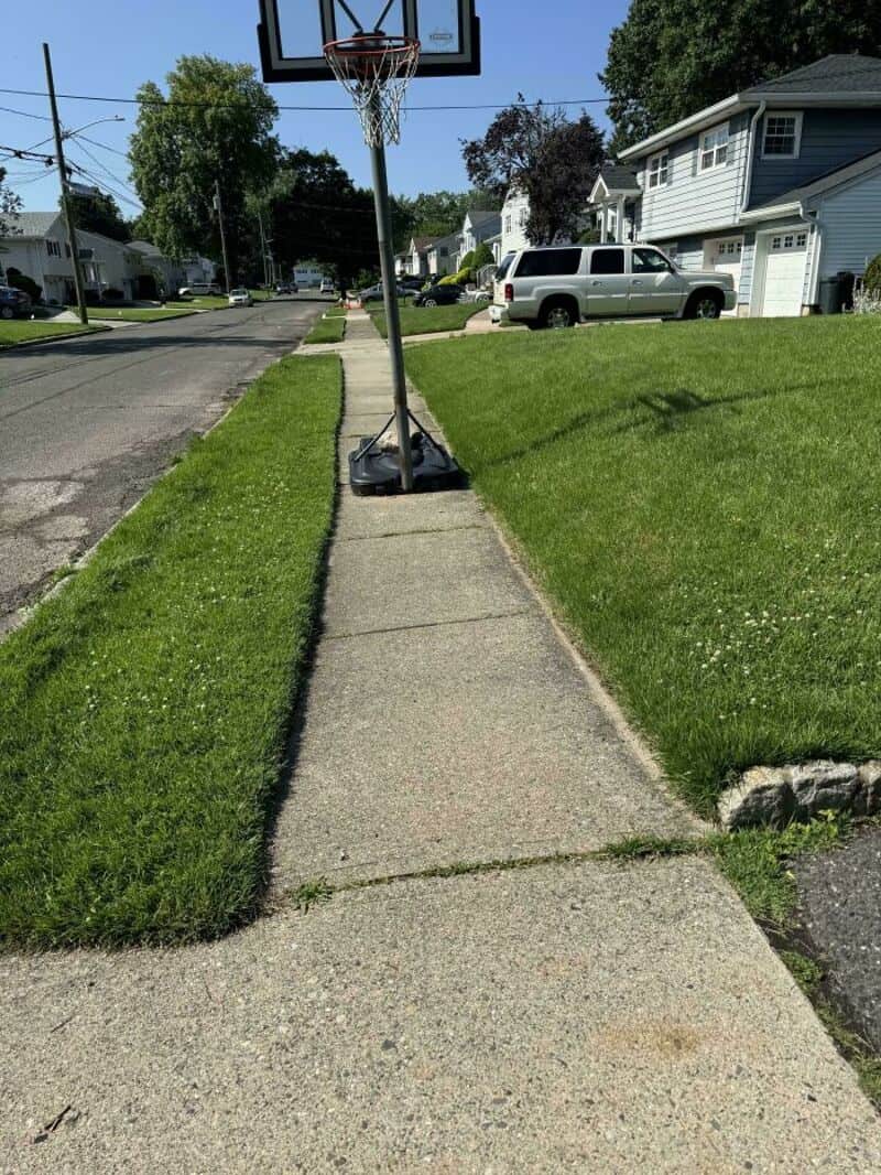 Basketball hoop on a sidewalk with a damaged base and a residential street background.