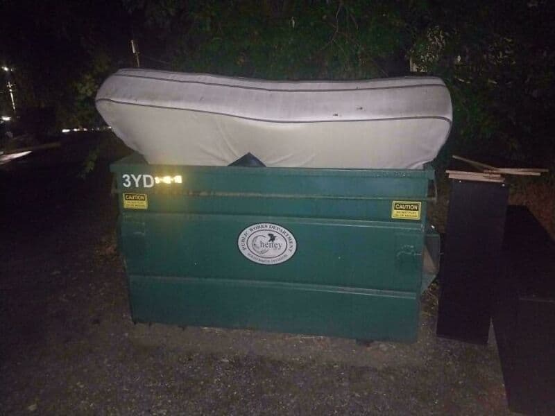 A boat is placed on top of a large green dumpster at night.