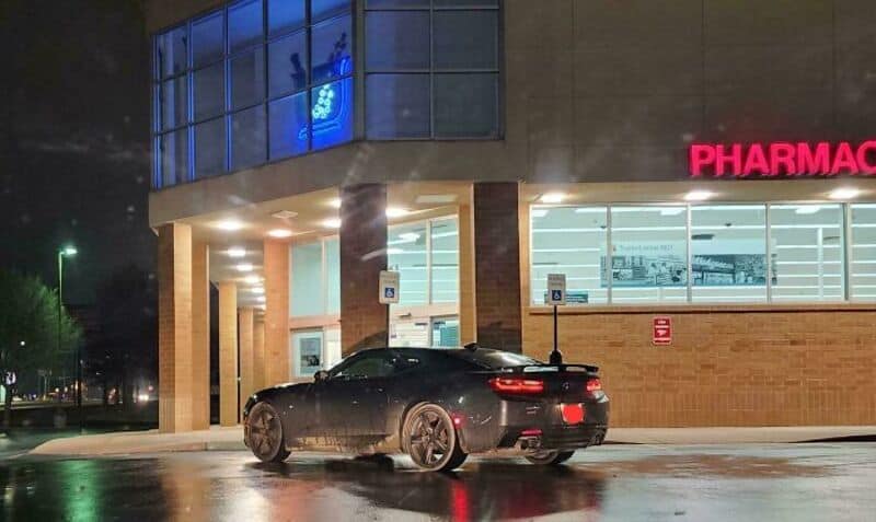 Black sports car parked outside a pharmacy at night with wet pavement.