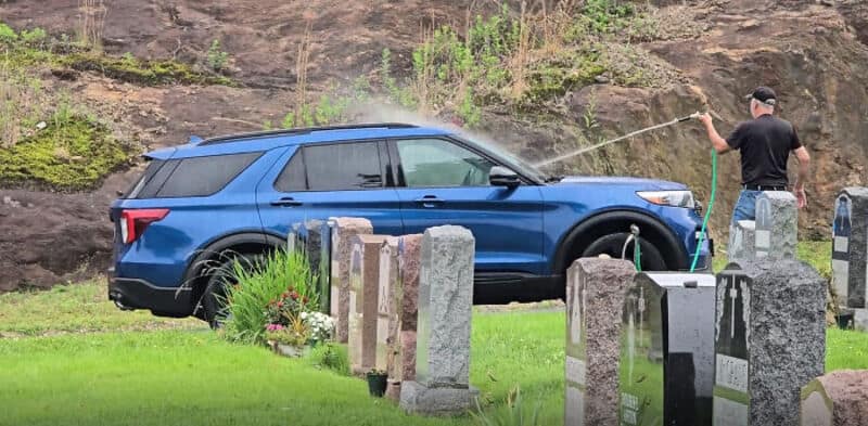 Man washing blue SUV in a graveyard with headstones and greenery.