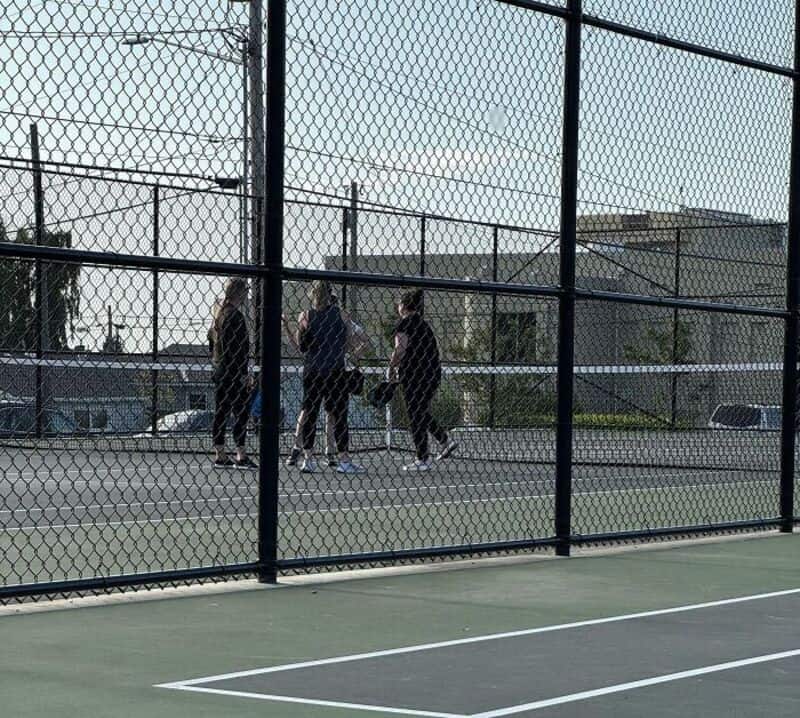 Four people playing basketball on an outdoor court, viewed through a chain-link fence.
