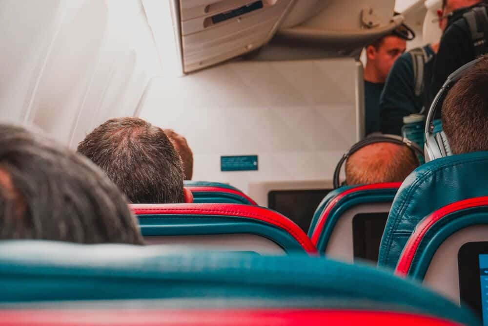 Passengers seated on an airplane, facing forward, with a flight attendant at the front.