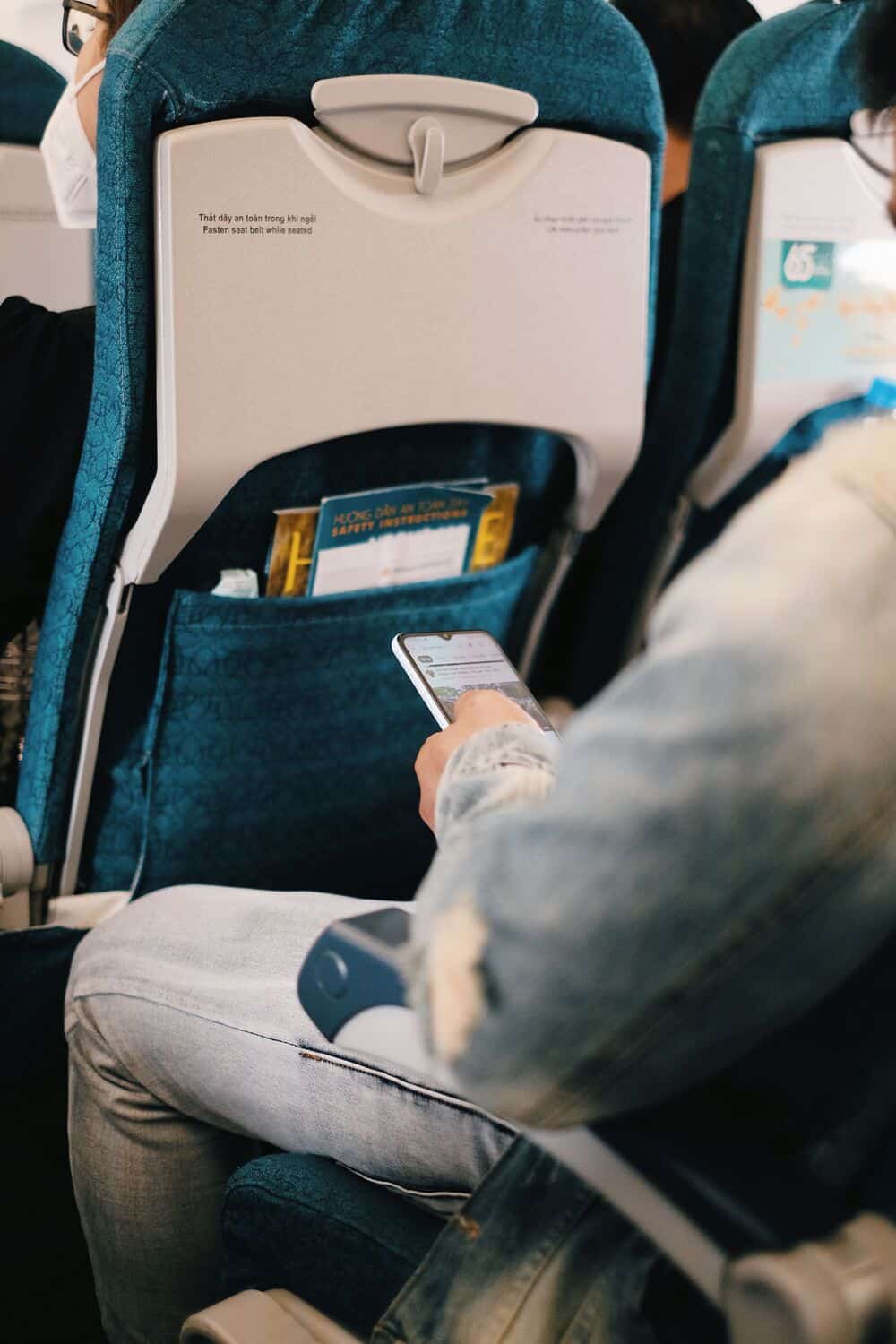 Passenger using smartphone on airplane seat with safety card in pocket.