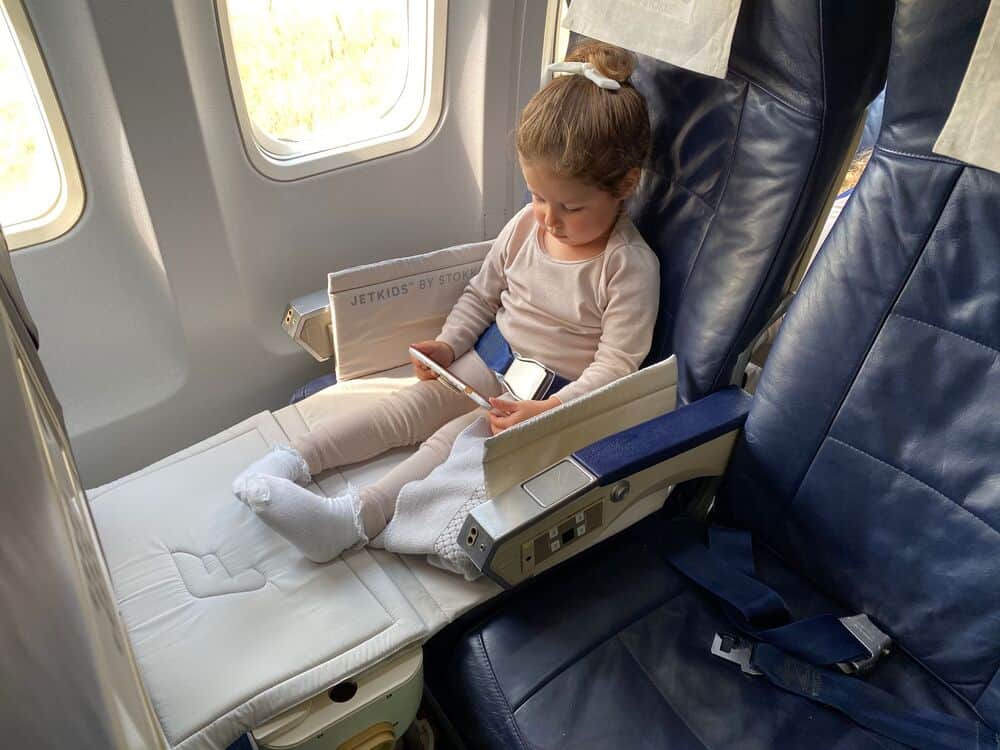 Young girl sitting in airplane seat, playing on a tablet, with window view.