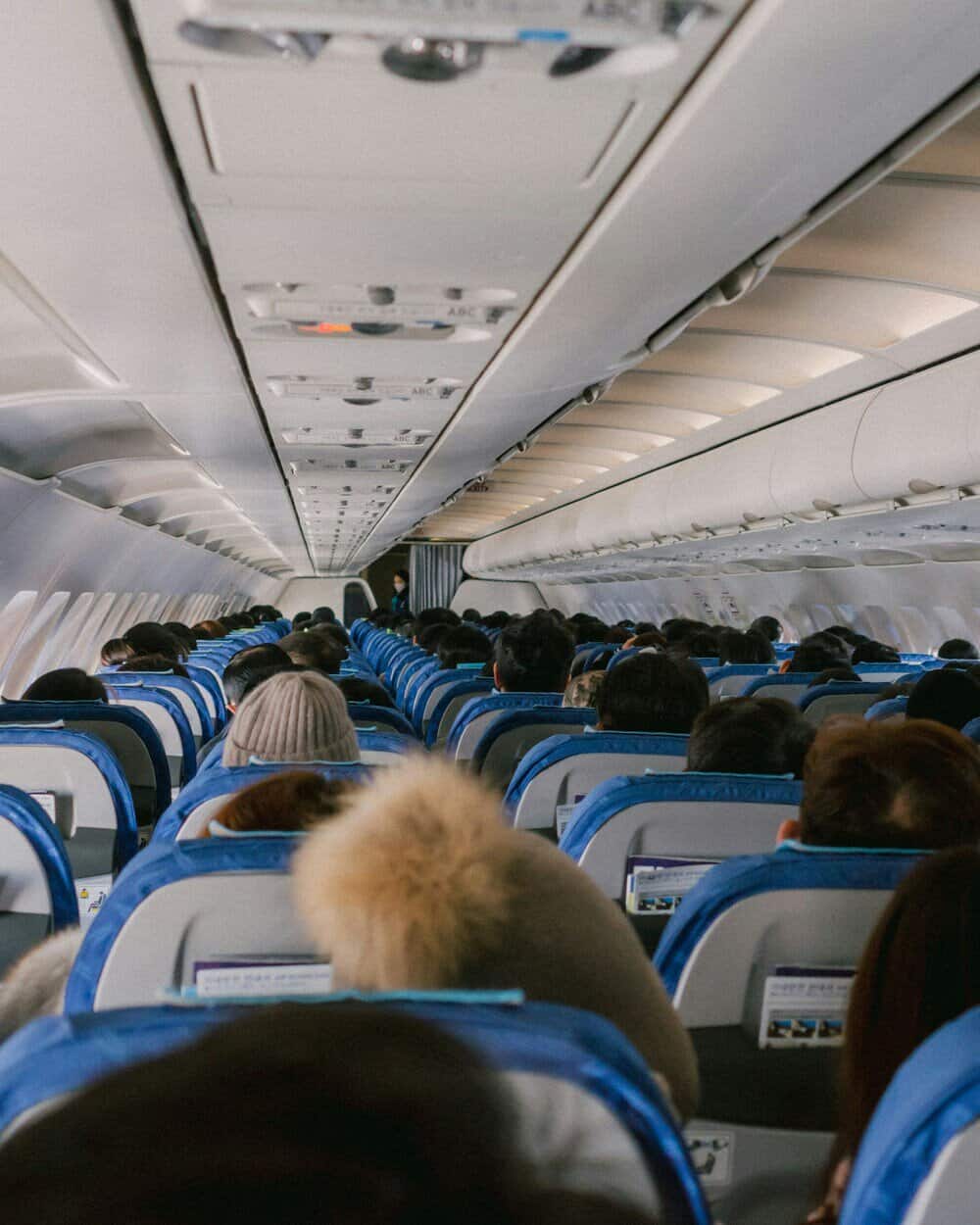 Passengers seated on an airplane, some using in-flight screens, aisle view.