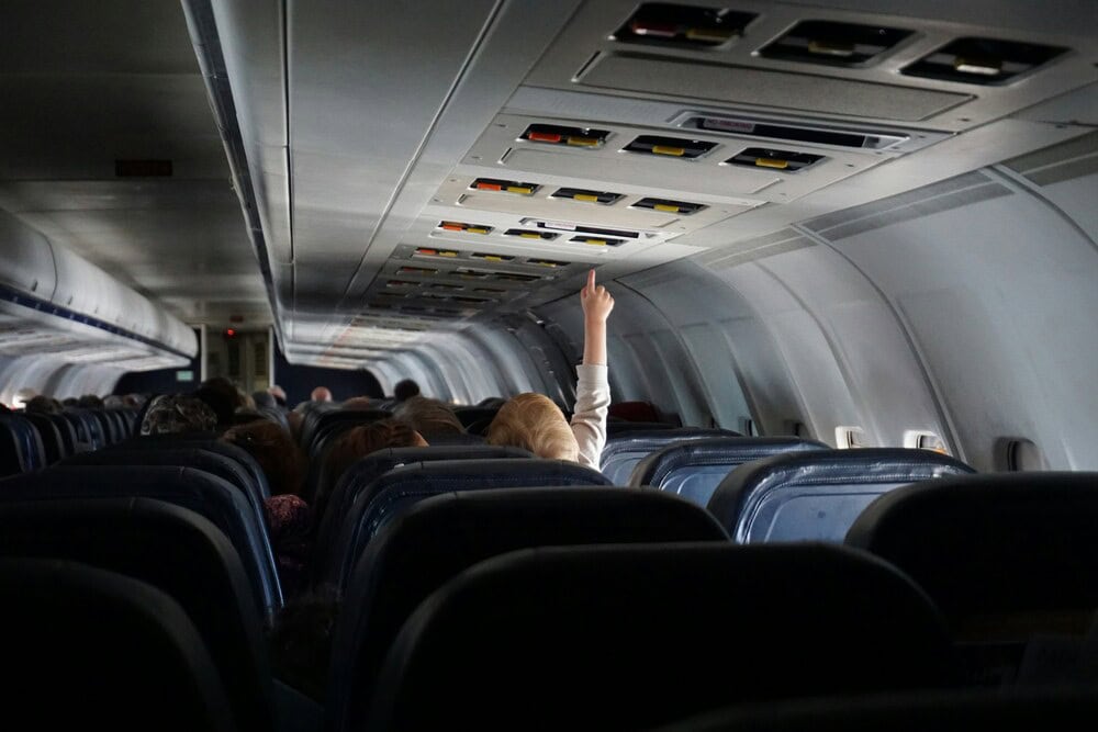 Passenger raising hand in airplane cabin, many seats and overhead compartments visible.