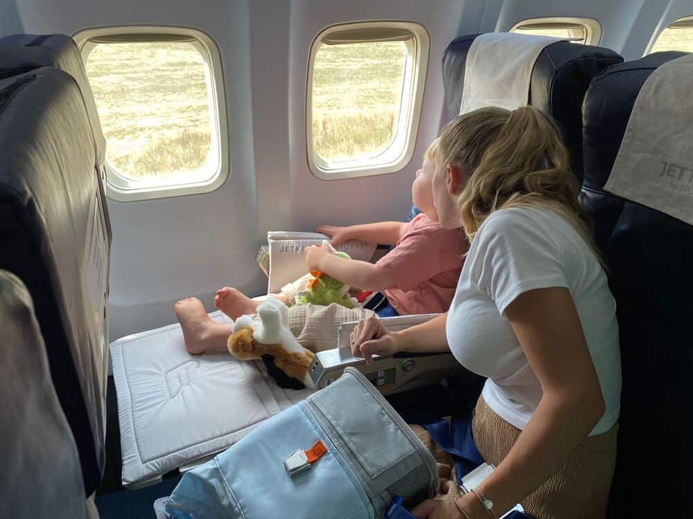 Two children sitting by airplane window, looking outside, with toys and snacks.