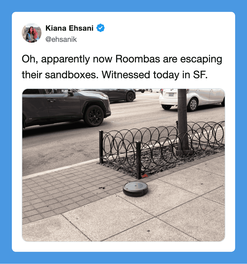 Roomba on sidewalk near a small fenced area with a tree, cars on street in background.
